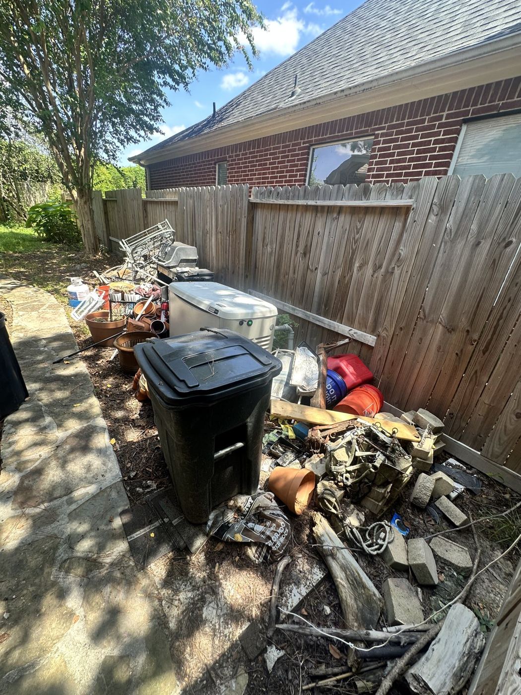 Side yard beside a brick house with a pile of old metal furniture, pots and bins